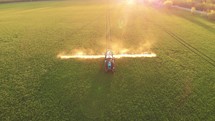 Aerial footage. Pesticide Sprayer Tractor working on a large green field at sunset. Aerial shot following on the side a tractor spraying wheat field against diseases. Farmer spraying soybean fields.