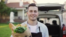 Close up handsome smiling and looking at the camera courier man going from the car to house and holding paper bag with vegetables outdoors. Worker grocery, online order, fast door to door delivery.