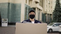 Delivery man in uniform, mask and gloves carrying big cardboard box parcel outdoor.