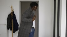 Black man in front home door putting on trendy coat, leaving house.