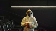 A man in white protection costume and respirator came alone to the cinema with bucket of popcorn, slow motion.