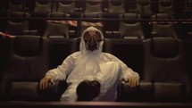 Portrait of a man in white protection costume and respirator sitting alone at the cinema, front view.