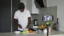 Young African American man cutting fresh cabbage at kitchen table while cooking vegetable salad.