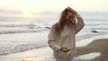 Beautiful young woman spinning around on ocean beach at sunset, filming herself with her phone.