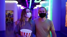 Happy young caucasian couple in mask go to the cinema, walking by hall with bucket of popcorn.