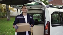 Handsome smiling man in uniform taking box out of trunk and looking at the camera. Parcel delivery, hand trolley.