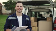 Happy smiling delivery service worker makes notes on documents and standing on the street near the minivan. Close up footage.