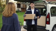 Woman meets a handsome friendly delivery man and takes a parcel box beside delivery van. Courier making notes about the parcel. Concept of courier, home delivery, online shopping.