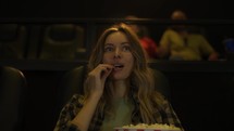 Excited woman sitting in armchair watching a movie at the cinema, eating popcorn.