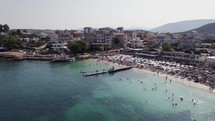 Aerial View Of Busy Crowded Plazhi Ksamil 9 Beach With Mini Dock, Albania. Dolly Establishing Shot
