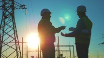 Silhouette of engineers looks at the construction of high-voltage power. Teams engineer looking discussing plan. Two engineer standing on field with electricity towers at sunset.