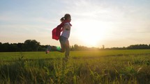 Girl Plays Superhero. Happy girl are playing superhero. Kid run across green field in red cloak at sunset time. Pretty child superhero hero in red cloak in nature. Girl power concept.