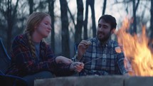Young man and woman sit beside a fire and make s’mores 