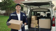 Happy smiling delivery service worker in uniform, cap and gloves holding paper box and standing near the minivan with opened trunk. Close up footage outdoor.