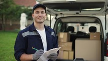 Happy smiling delivery service worker in uniform, cap and gloves makes notes on documents and standing on the street near the minivan. Close up footage.