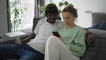 Happy biracial couple relaxing on sofa with smartphone.
