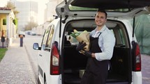 Happy delivery worker holding paper bag with food looking at the camera and smiling. Close up portrait of positive young man courier person in black apron and gloves.