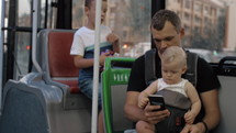 Dad riding in the bus with elder son and one year old baby daughter. Everyone watching fathers smart phone