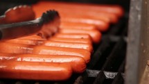Tongs moving hot dogs on a grill.