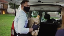 Female customer is getting paper bag of organic groceries from male courier in a mask, apron and gloves.