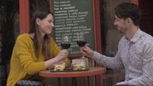 A young couple sitting at a small table of cafe terrace, drinking red wine from big glasses and trying cheese from a cheese plate