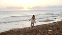 Happy young woman with long blonde hair in white suit running along ocean beach at sunset.