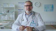 Mid-Aged Doctor Sitting in Clinic and Giving Online Consultation
