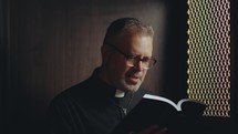 Catholic priest wearing traditional cassock and glasses sitting inside confessional and reading Bible to penitent
