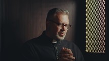 Mature Catholic priest wearing black cassock sitting inside of confessional booth, holding hands together and praying to God
