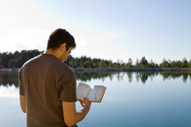Man reading Bible while standing by a lake.