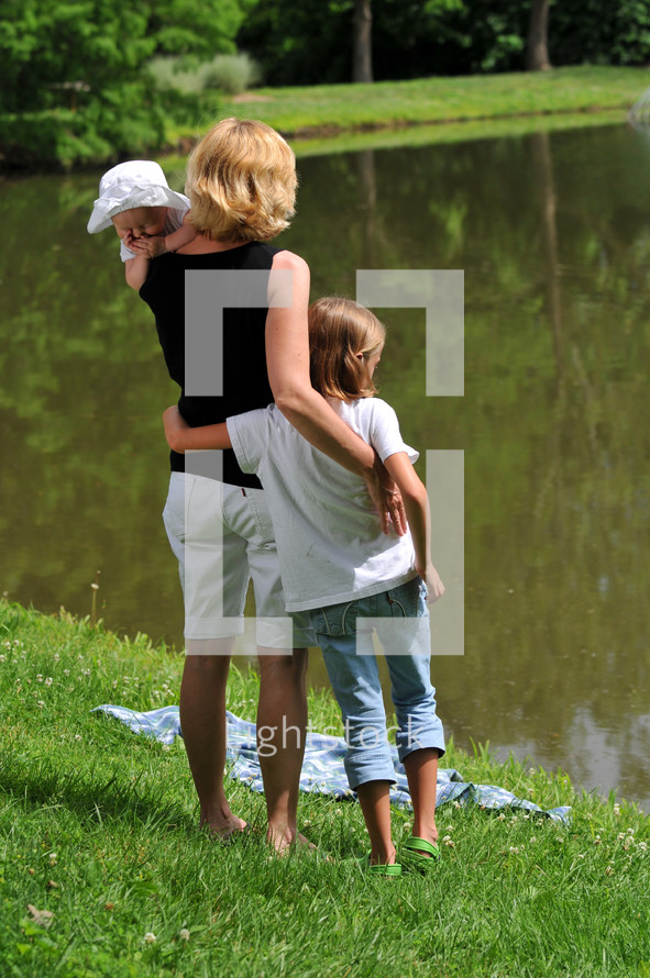 Woman with infant in her arms and a arm around a young girl, standing by a lake