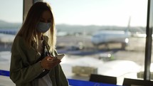 Blonde woman in medical mask uses phone walking through airport terminal.