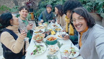 Company of cheerful friends sitting together at table with food and drinks, looking at camera, smiling and talking while video calling or taking selfie on outdoor dinner party
