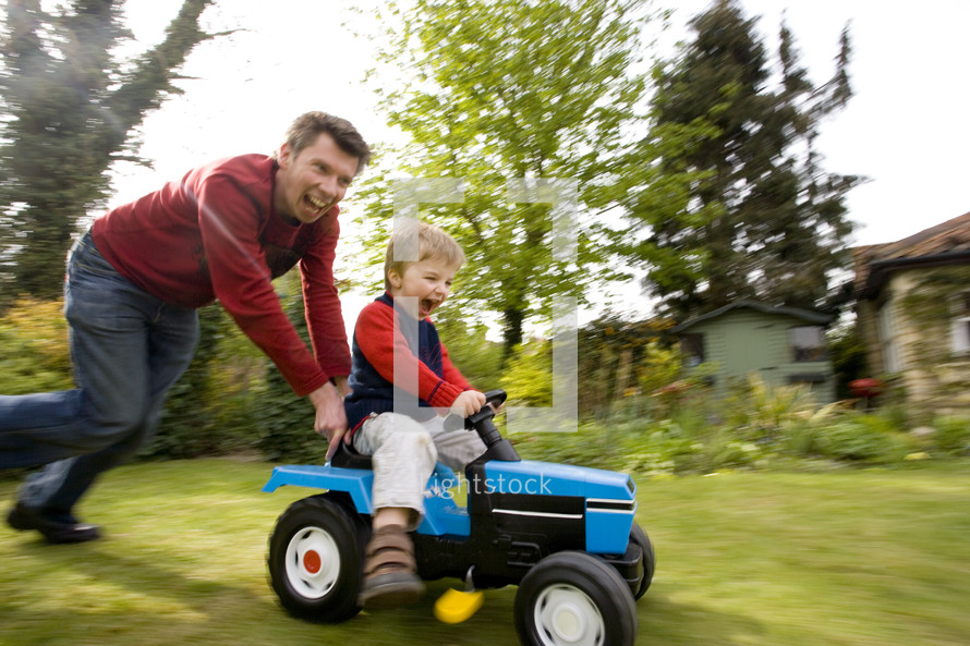 father pushing his son on a toy tractor in their back garden themes of exhilaration family bonding