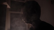 Troubled young man, teenage boy smoking drugs or cigarette, getting high, in dark, smoky room in cinematic slow motion.