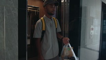 Young male courier with backpack leaving elevator, walking through corridor in residential building with bag, delivering food and drinks to customer
