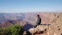 Stunning nature of great Grand Canyon in National Park. Male tourist sitted at the edge at top of observation deck and enjoys views of red rocks and canyon gorges 
