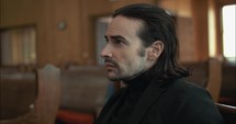 Young, emotional, anxious, and stressed man with long hair and black suit sitting in old church in worship and praying.