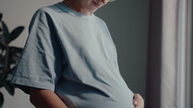 Happy pregnant woman standing at home, stroking her belly with love, looking through the window, smiling. Tilt up shot
