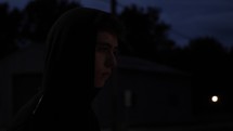 Young, sad looking and depressed young man or teenage boy walking alone at night in small town while wearing a hood in cinematic slow motion.