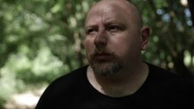Middle aged man in black shirt with beard in nature walking, hiking, meditating, praying contemplating in green, wooded area in trees with sunlight shining in cinematic, slow motion.