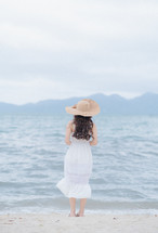 a woman in a white dress and sunhat on a shore 
