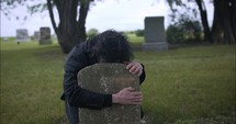 Sad, grieving man embracing family tombstone in cemetery crying in cinematic slow motion. 