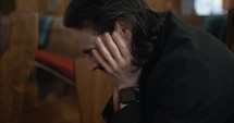 Young, emotional, anxious, and stressed man with long hair and black suit sitting in old church in worship and praying.