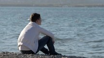 Guy sitting on a rocky shore, looking out at a body of water. 