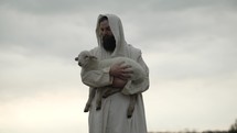 Jesus Christ, the good shepherd of Psalm 23, holding a baby lamb or sheep in his arms while wearing a white, tattered tunic and hood outside in front of cloudy sky during sunset.
