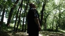 A Christian man in black shirt with beard in nature walking, hiking, meditating, praying contemplating in green, wooded area in trees with sunlight shining in cinematic, slow motion.
