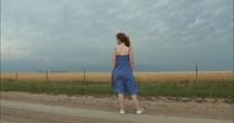 A young woman in a blue dress standing outside on a dirt road in farm land under a stormy, moody, cloudy sky as distant storm approaches. 