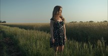 Young woman in flower dress watching summer sunset standing in green grass field. 