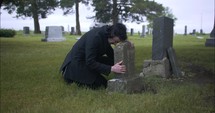 Young, sad man in black suit kneeling, crying, hugging tombstone in cemetery in cinematic slow motion.